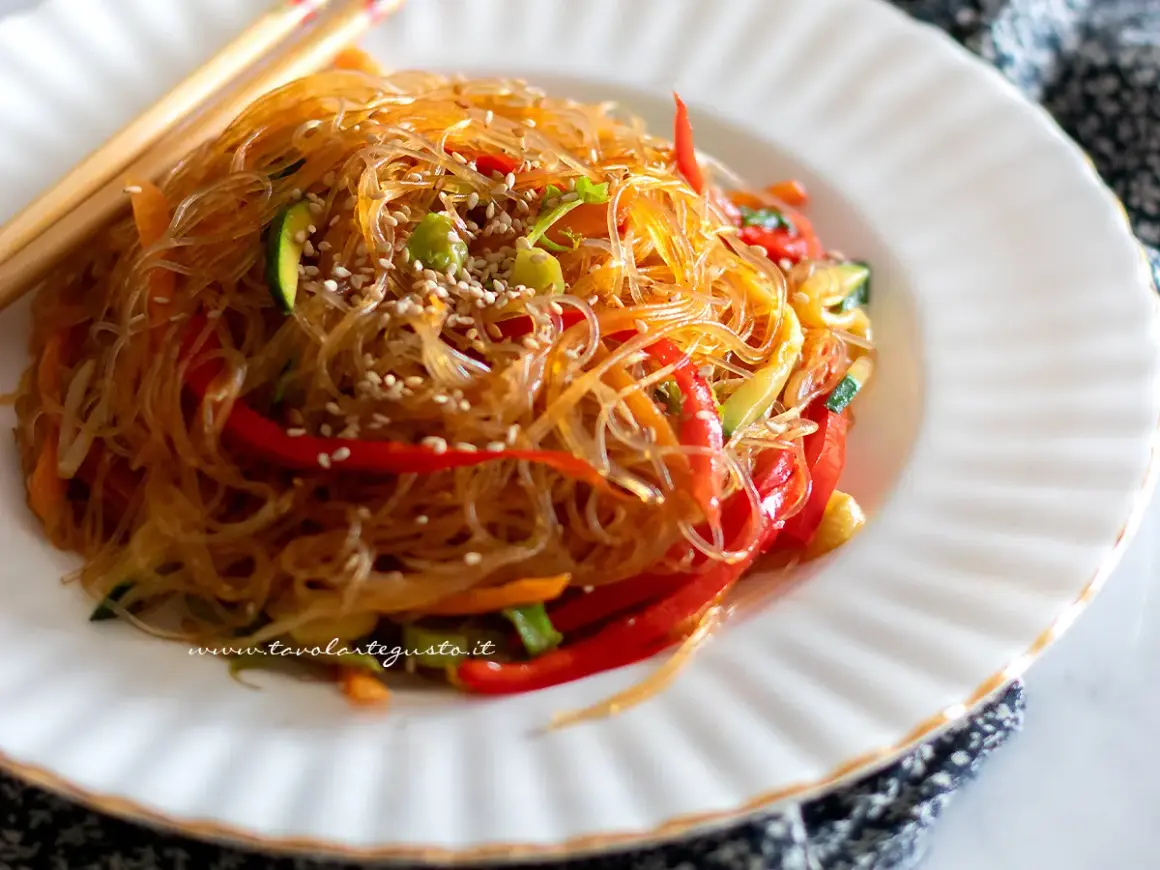 Un piatto invitante di spaghetti di soia con verdure colorate, peperoni rossi, zucchine e carote, guarnito con semi di sesamo.