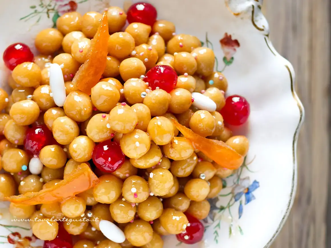 Struffoli ricetta napoletana: palline dorate glassate, ciliegie rosse, canditi e confettini in un piatto decorato.