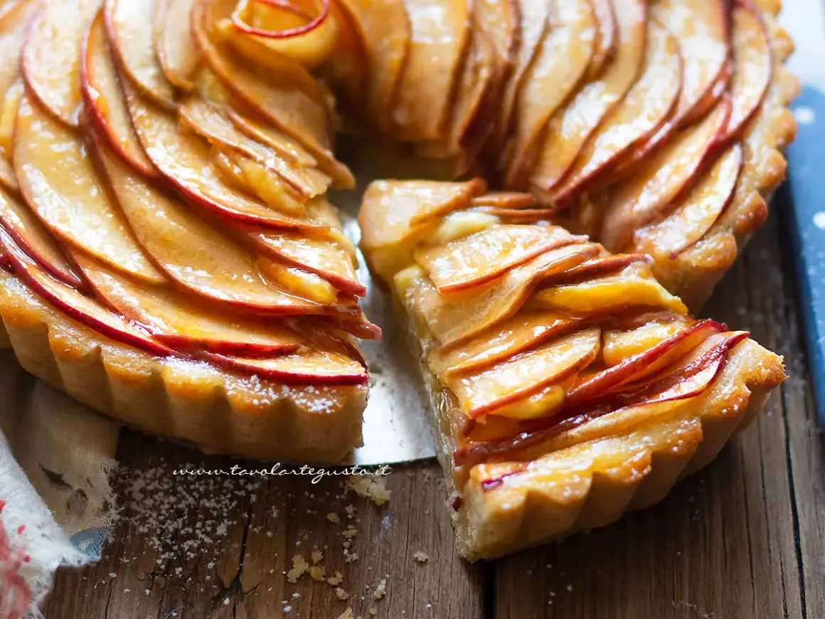 Una deliziosa crostata di mele, con fette di mela lucide disposte a spirale su una base dorata. Una fetta è stata tagliata, rivelando la morbidezza del ripieno.