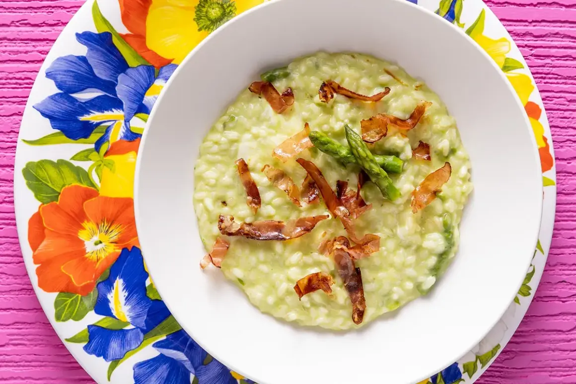 Un cremoso risotto asparagi e speck, guarnito con punte di asparagi freschi e croccanti listarelle di speck, servito su un piatto fiorito.