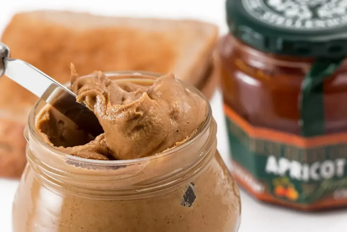 Un barattolo di burro di arachidi cremoso, pronto per essere spalmato su una fetta di pane tostato. Accanto, una marmellata di albicocche.