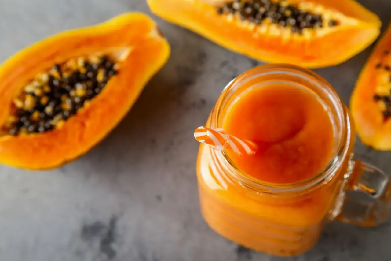 Frullato di papaya in un bicchiere con cannuccia a righe, accanto a met&agrave; papaya. Un aiuto naturale per chi vuole imparare come sgonfiare la pancia.