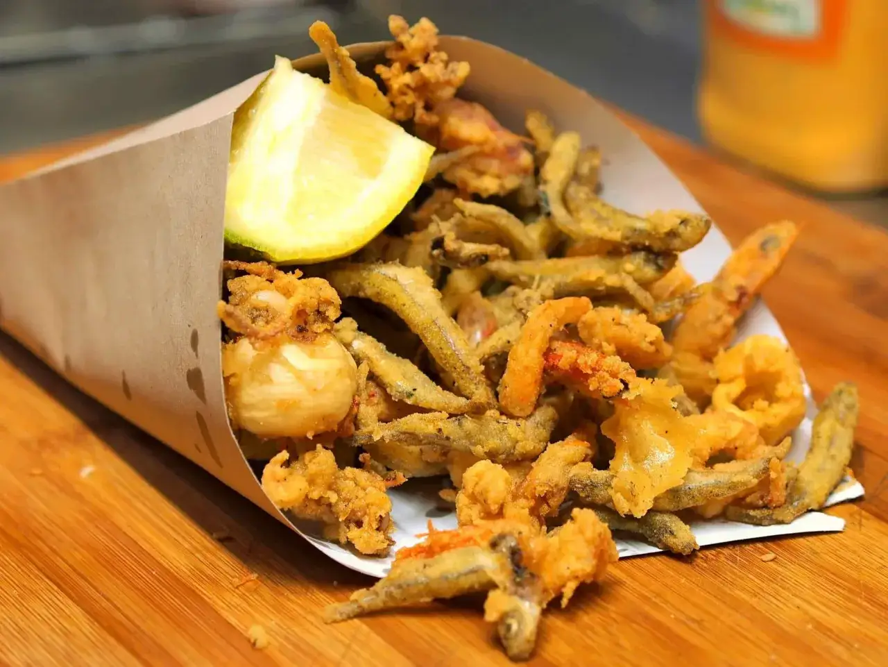 Un cono di carta marrone trabocca di una deliziosa frittura di pesce croccante, con calamari, gamberetti e pesciolini, guarnito con uno spicchio di limone.