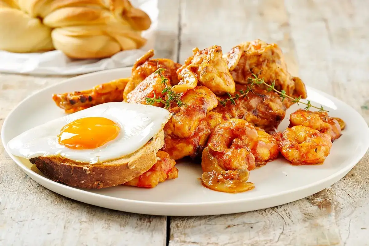 Piatto di pollo alla marengo con gamberi, uovo fritto su pane tostato e un contorno di pane intrecciato.