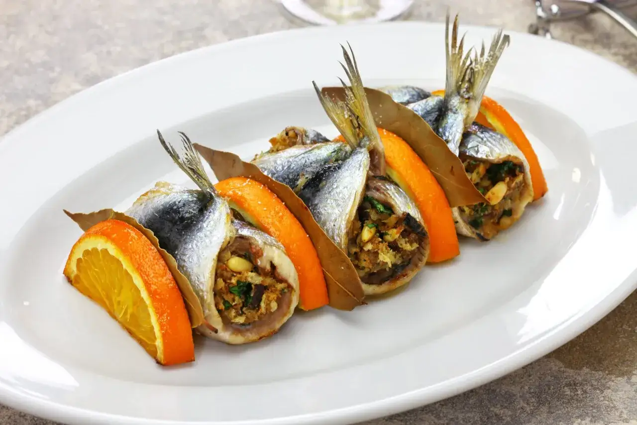 Sarde a beccafico ripiene, guarnite con fette d'arancia e foglie di alloro, presentate su un piatto bianco.
