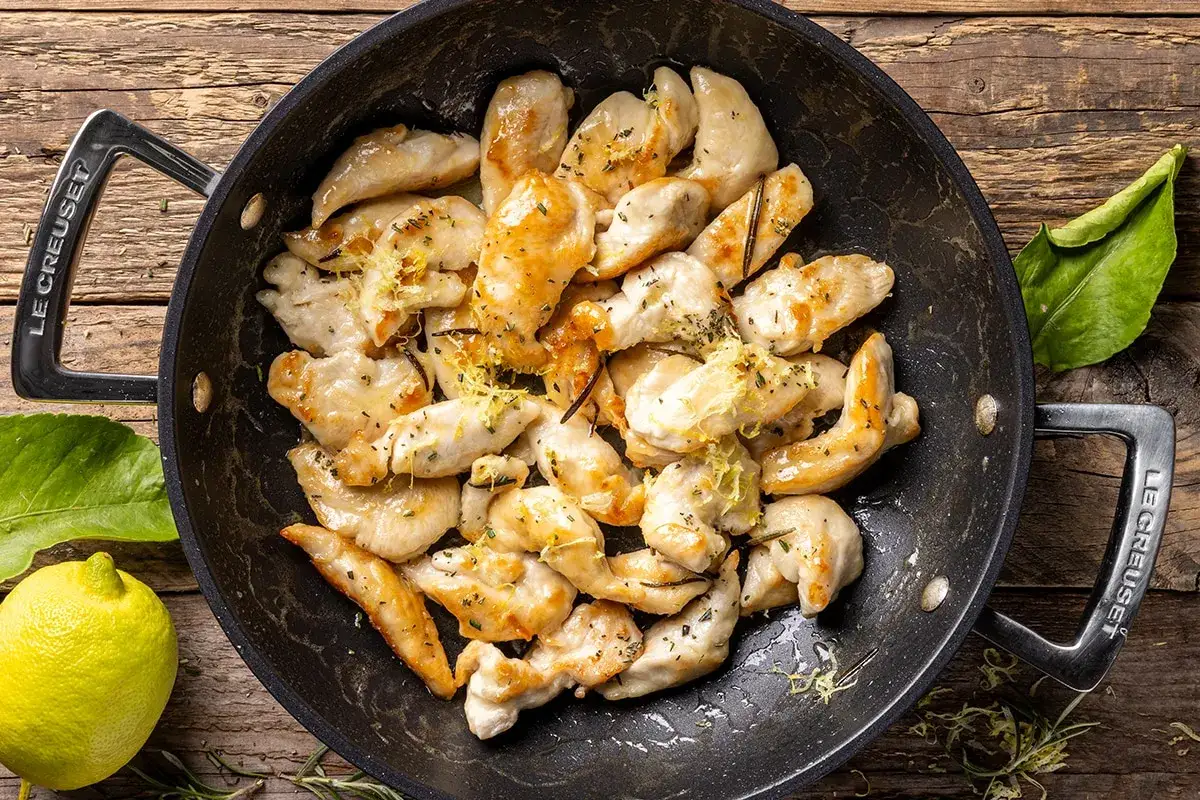Straccetti di pollo dorati in padella, conditi con scorza di limone e erbe aromatiche, pronti per essere gustati.