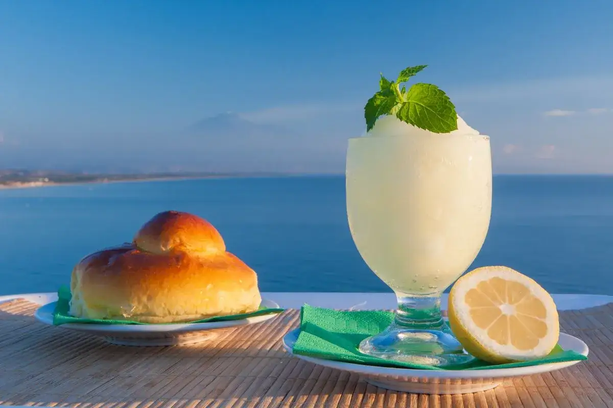 Una brioche e una coppa di granita al limone, guarnita con menta, con il mare e un vulcano sullo sfondo.