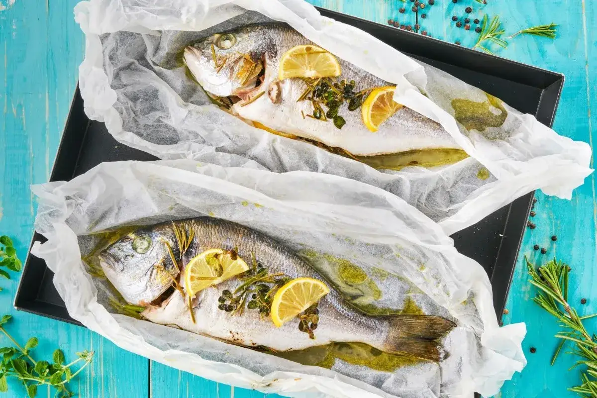 Due orate al cartoccio, pronte per essere infornate, con fette di limone e erbe aromatiche.