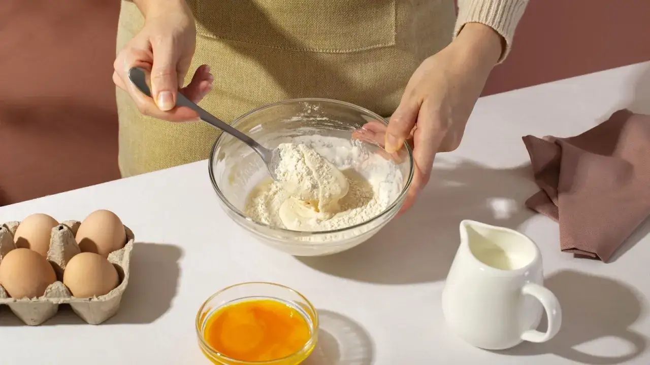 Mani che mescolano farina e ingredienti in una ciotola, forse per preparare una torta con bicarbonato al posto del lievito. Uova, tuorlo e latte a portata di mano.