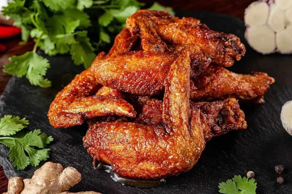 Alette di pollo dorate e croccanti, servite su una pietra scura con coriandolo, zenzero e aglio.