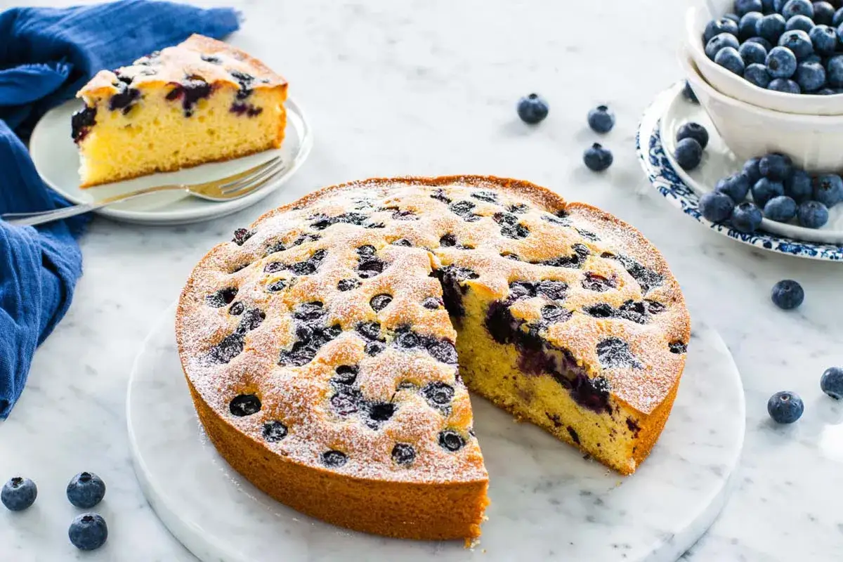 Una deliziosa torta 7 vasetti ai mirtilli, cosparsa di zucchero a velo, con una fetta tagliata e mirtilli freschi sparsi.