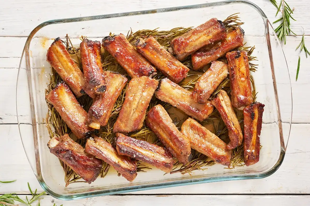 Succulente costine al forno, dorate e profumate al rosmarino, servite in una pirofila di vetro.