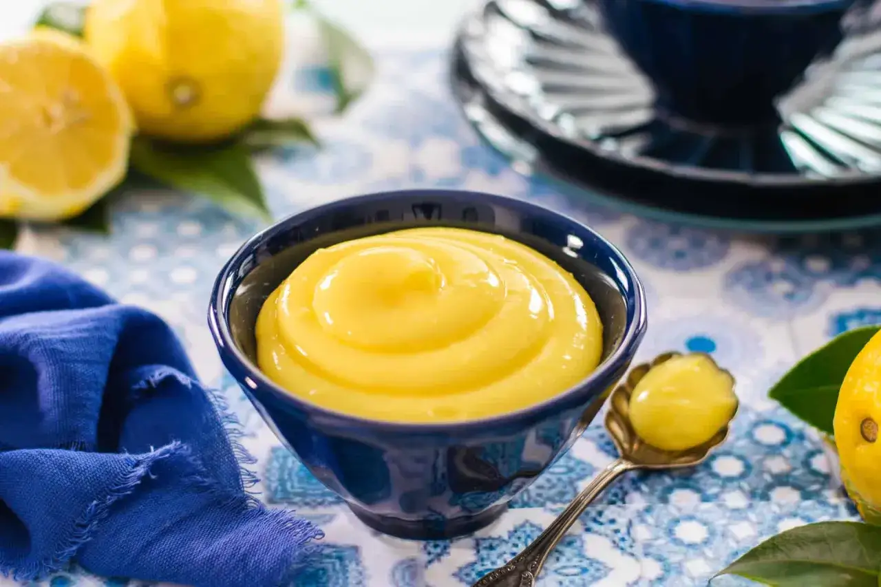 Una ciotola blu piena di deliziosa crema al limone, con limoni freschi e un tovagliolo blu sullo sfondo.