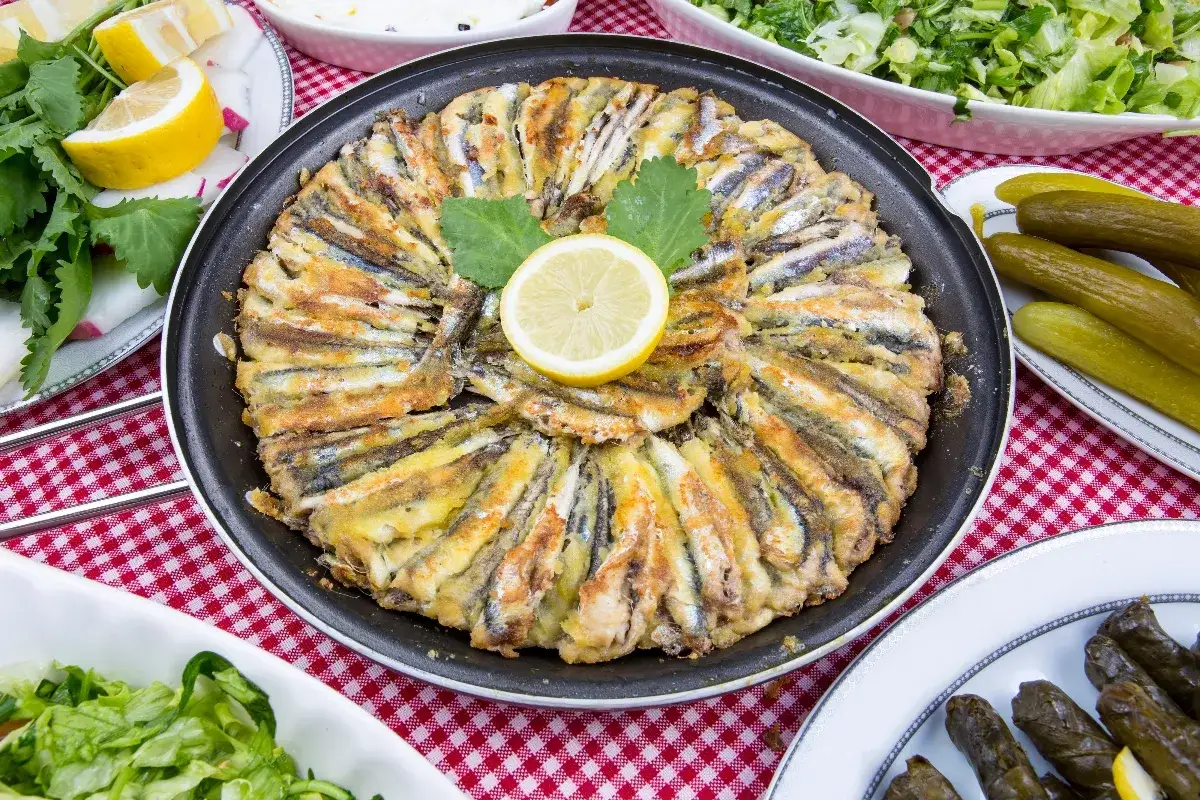 Alici in tortiera dorate e croccanti, disposte a raggiera in una padella, con una fetta di limone al centro. Accanto, insalata fresca e cetriolini.