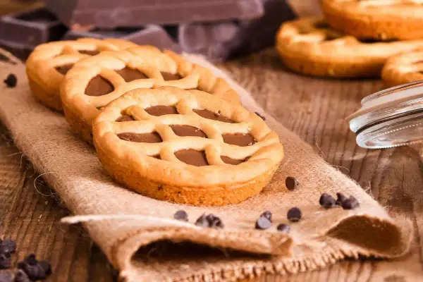 Deliziose crostatine al cioccolato con una griglia di pasta frolla dorata, su un sacco di juta, con gocce di cioccolato sparse.
