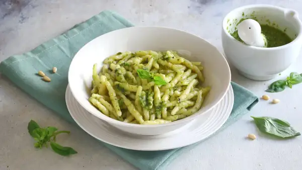 Un piatto invitante di pasta al pesto, condita con basilico fresco e pinoli. Accanto, un mortaio con altro pesto fatto in casa.
