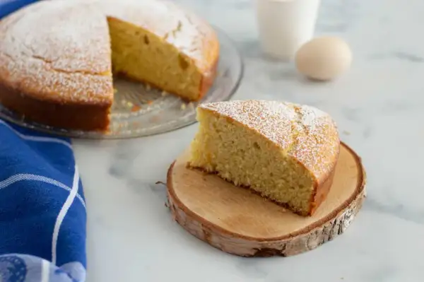 Una fetta di torta 7 vasetti, soffice e cosparsa di zucchero a velo, su un tagliere di legno. Sullo sfondo, la torta intera e un uovo.