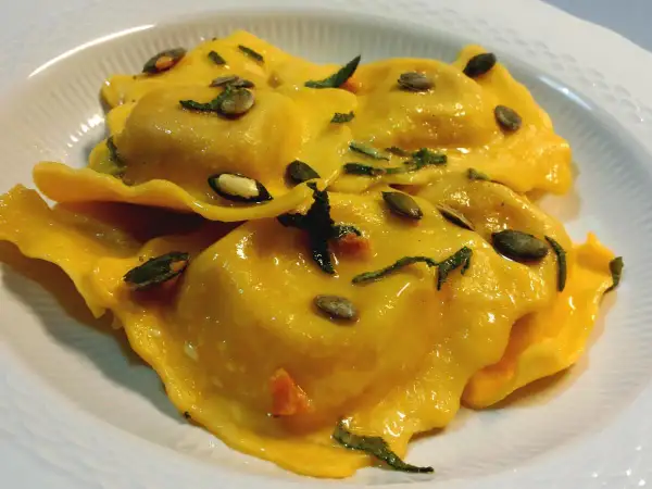 Tortelli di zucca dorati, conditi con erbe aromatiche e semi di zucca tostati, serviti in un piatto bianco.