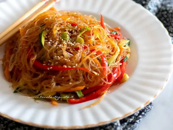 Un piatto invitante di spaghetti di soia con verdure colorate, peperoni rossi, zucchine e carote, guarnito con semi di sesamo.