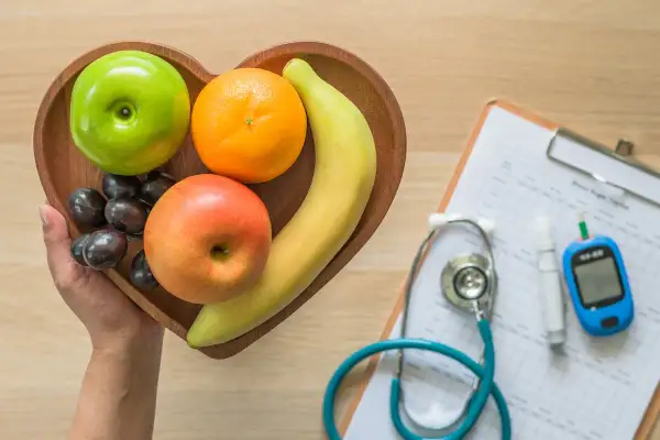 Frutta fresca in un vassoio a forma di cuore, accanto a uno stetoscopio e un glucometro, per capire come abbassare la glicemia con una dieta sana.
