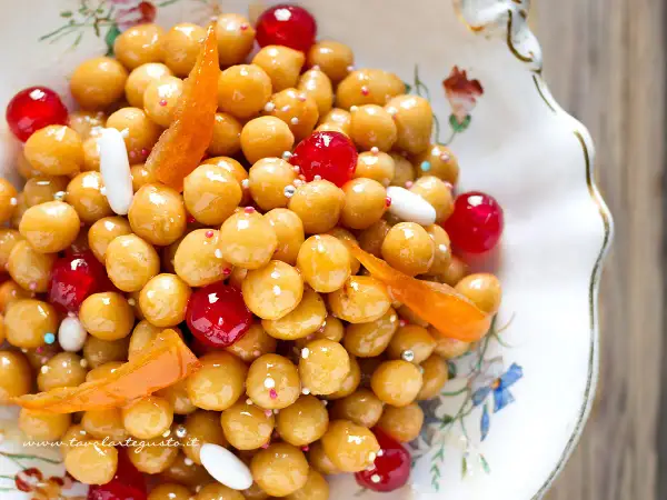 Struffoli ricetta napoletana: palline dorate glassate, ciliegie rosse, canditi e confettini in un piatto decorato.