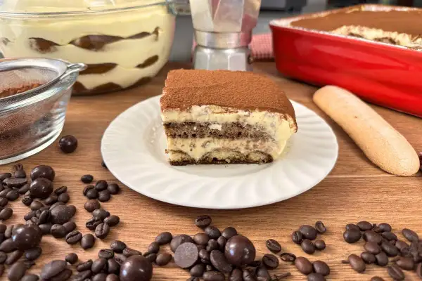 Una fetta di tiramisù con uova pastorizzate, cosparso di cacao, su un piatto bianco, circondato da chicchi di caffè e cioccolatini.