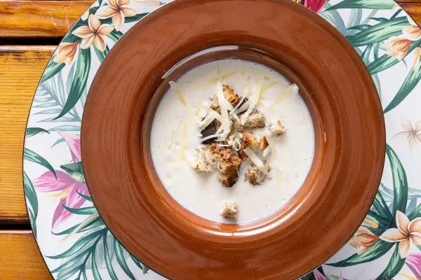 Una cremosa zuppa di cipolle, guarnita con crostini dorati e formaggio grattugiato, servita in una ciotola marrone su un piatto floreale.