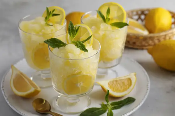 Tre bicchieri di rinfrescante granita al limone, guarniti con fettine di limone e menta. Limoni freschi e un cucchiaino dorato completano la scena.
