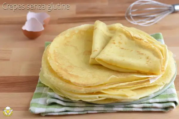 Pila di crepes senza glutine dorate e soffici, pronte per essere farcite. Sullo sfondo, gusci d'uovo e una frusta.